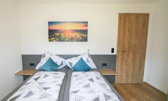 A modern bedroom with two single beds and colorful cushions. On the wall hangs a picture of a sunset over a meadow.