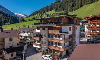 A modern hotel with wooden cladding in a picturesque mountain landscape. In the background, green meadows and snow-capped mountains can be seen.