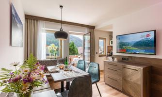 A bright and modern living kitchen with a dining table and chairs. A television on the wall and large windows provide a view of the mountains.
