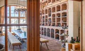 An elegant wine cellar with wooden shelves full of wine bottles. In the background, there is a long table with chairs and a large window front.