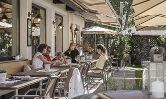 An inviting outdoor café with several guests sitting at the table. Umbrellas provide shade, and a green environment creates a relaxed atmosphere.