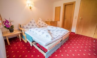 A cozy bedroom with a double bed and red carpet.  
On the wall, there is a light source and a small table with a flower.