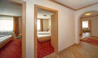A bright room with a double bed and another bedroom in the background. The floor is tiled, and large windows provide pleasant lighting.