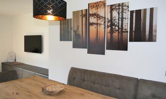 A modern dining room view with a large wooden table and a comfortable bench. Pictures of trees at sunset hang on the wall, and a stylish lamp provides lighting.
