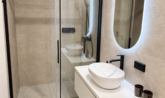 A modern bathroom with a glass shower and an elegant sink arrangement. The walls are decorated with light tiles, and a round mirror with lighting creates a stylish ambiance.