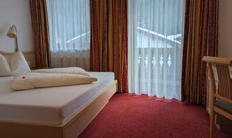 A cozy bedroom with a red carpet and large windows. The bed has a simple setup with pillows and a bright, inviting atmosphere.
