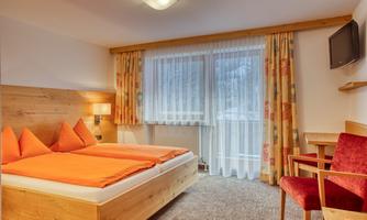 A cozy bedroom with a double bed and orange pillows. Large windows with curtains let in plenty of light, and there is a chair as well as a television.
