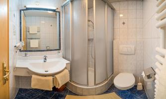 A small bathroom with a shower, a sink, and a toilet. The walls are light-tiled, and there is a large wall mirror.
