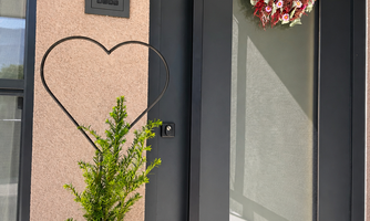 A modern front door with a large glass frame and a decorative flower wreath. Next to the door stands a green plant and a heart shape.