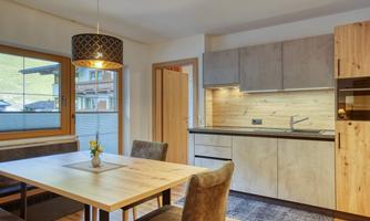 A modern kitchen with wooden cabinets and a dining table. There are large windows that let natural light into the room.
