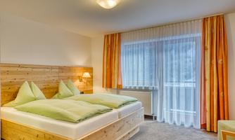 A cozy bedroom with two single beds and green pillows. Large windows with curtains allow daylight to come in.