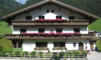 A traditional alpine house with balconies decorated with colorful flowers. In the background, green mountains and a blue sky can be seen.