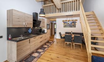 A modern kitchen with wooden cabinets and a dining table is visible in the foreground. A wooden staircase leads to an upper area.