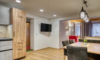 A modern living room with an open kitchen and a dining area. There is hardwood flooring, a TV on the wall, and plenty of natural light.