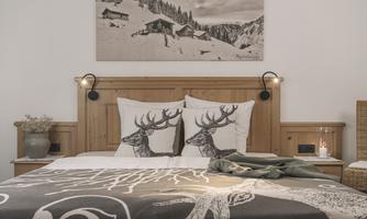 A cozy bedroom with wooden details and a large bed. The pillows feature deer motifs, and above the bed hangs a wintry picture of mountain landscapes.