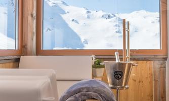 A cozy room with large windows that offer a view of snow-covered mountains. In the room, there is a bucket and a blanket inviting relaxation.