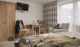 A cozy bedroom with a double bed and a small table. The walls are light-colored, and there is a television as well as a large window front with curtains.