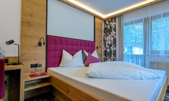 A modern bedroom with a large bed and a pink headboard. The room has large windows that let in plenty of daylight.