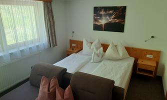 A cozy bedroom with two beds and a gray sofa. A large window provides bright light and a calm atmosphere.