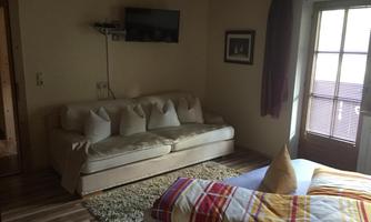 A cozy room with a sofa and a large bed. It has wooden flooring and windows with curtains.