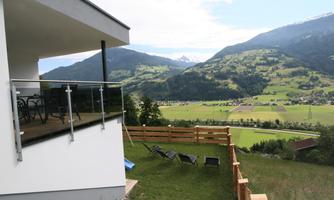 A modern terrace overlooking a green valley landscape with mountains in the background. The weather is clear and sunny.