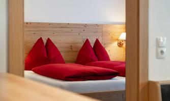 A cozy bedroom with a double bed and red pillows. The walls are made of wood, and a lamp provides soft light.