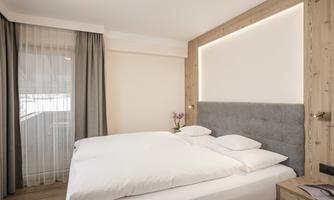 A modern bedroom with a cozy double bed and soft lighting.  
Elegant curtains hang in front of a window front, and a flower stands on the nightstand.