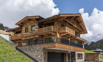A modern wooden house with a spacious balcony and stone facade. It is located in a picturesque mountain landscape under a clear sky.