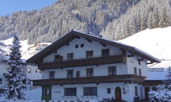 A cozy chalet in the snow, surrounded by a picturesque mountain panorama. The landscape is calm and wintry with snow-covered trees.
