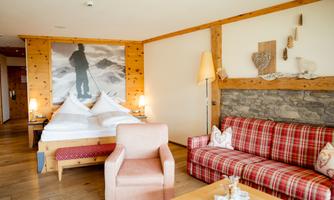 A cozy hotel room with wood paneling and a large bed. On the right side, there is a checkered sofa and a table with drinks.