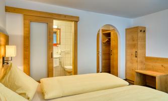 A cozy hotel room with two single beds and wooden furniture. In the background, a bathroom and a wardrobe are visible.