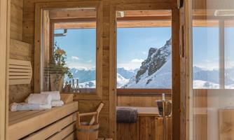 A cozy sauna with wooden interiors and windows that offer a view of snow-covered mountains. The atmosphere is relaxing and inviting.
