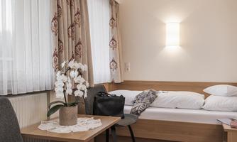 A modern hotel room with a single bed and a table. On the table is a vase with white orchids, and a black backpack is lying beside it.