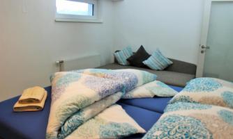 A cozy bedroom with a double bed and pillows. In the background, there is a sofa with colorful cushions.
