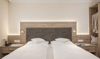 A modern bedroom with two single beds and a wooden wardrobe. The walls are painted light and there is soft lighting behind the headboard.