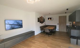 A modern living room with an open kitchen and a dining table. The walls are painted light and the floor is made of wood, creating a cozy atmosphere.