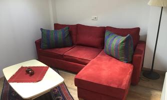 A red sofa with colorful cushions is in a bright room. Next to the sofa, there is a lamp and a small table with a red tablecloth.