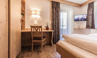 A cozy room with wooden details, a desk and a chair. The room features a window with curtains and a television on the wall.