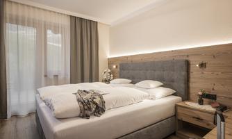 A modern bedroom with a large bed and warm wooden elements. The windows are decorated with light curtains, creating an inviting atmosphere.