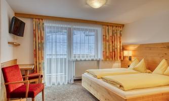A cozy room with two single beds and a wooden design. There is a television and a chair as well as large windows with curtains.