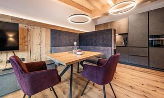 A modern dining room with a wooden floor and a stylish table. Two purple chairs and a kitchenette in the background complement the inviting space.