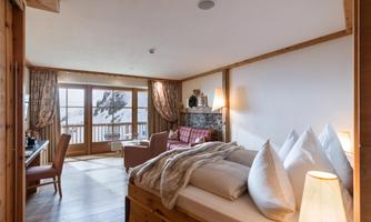 A cozy hotel room with wooden furniture and large windows. The view shows a snowy landscape, and the room is furnished with a bed, an armchair, and a table.