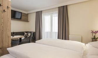 A comfortable room with two single beds and a television on the wall. The room features large windows with curtains and a cozy seating area.