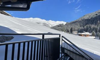 A snow-covered slope with mountains in the background. The sky is clear and blue, and some houses are visible in the valley.