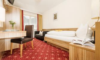 A bright room with wooden furniture and red carpet flooring. There is a single bed, a desk, and a cozy seating area.