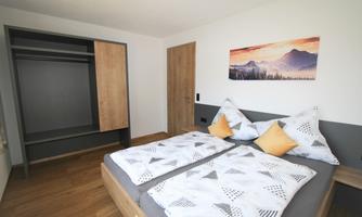 A modern bedroom with two beds and colorful pillows. In the background, there is a wardrobe and a picture of a mountain landscape.