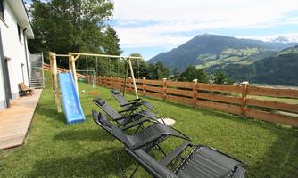 A spacious garden with sun loungers, a swing, and a slide. In the background, green hills and mountains are visible.