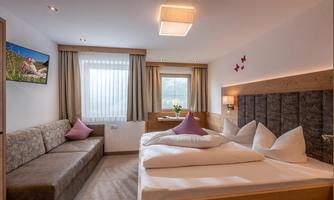 A modern bedroom with a double bed and a cozy sofa. The room is bright and welcoming, with large windows and a decorative wall.