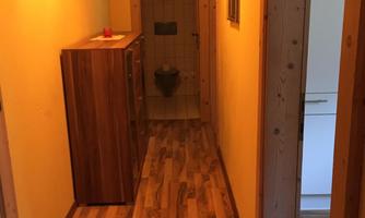 A narrow hallway with warm, yellow light. Jackets hang on the wall, and a dresser is on the left side.