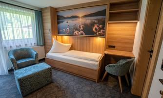 A modern room with a single bed and wooden paneling. It includes a comfortable chair, a footstool, and a view of the Tux Alps featuring a great picture by our local photographer Pfister Tom.